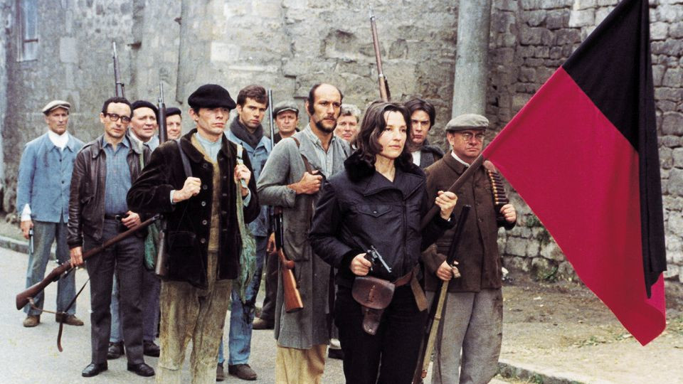 Filmstil, das eine Gruppe Menschen zeigt, die bewaffnet sind und eine rot schwarze Fahne vor sich her tragen.