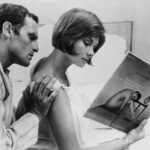 Filmstill aus Eine verheiratete Frau (Une femme mariée) 1964. Ein Mann im Schlafanzug berührt eine Frau, auf einem Bett sitzend mit einem Katalog/Bildband in der Hand, von hinten an der Schulter.