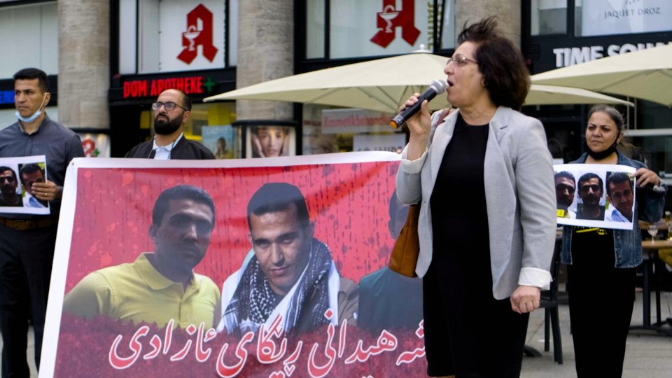Filmstill aus Mina, Frau mit Mikrofon steht vor Plakat