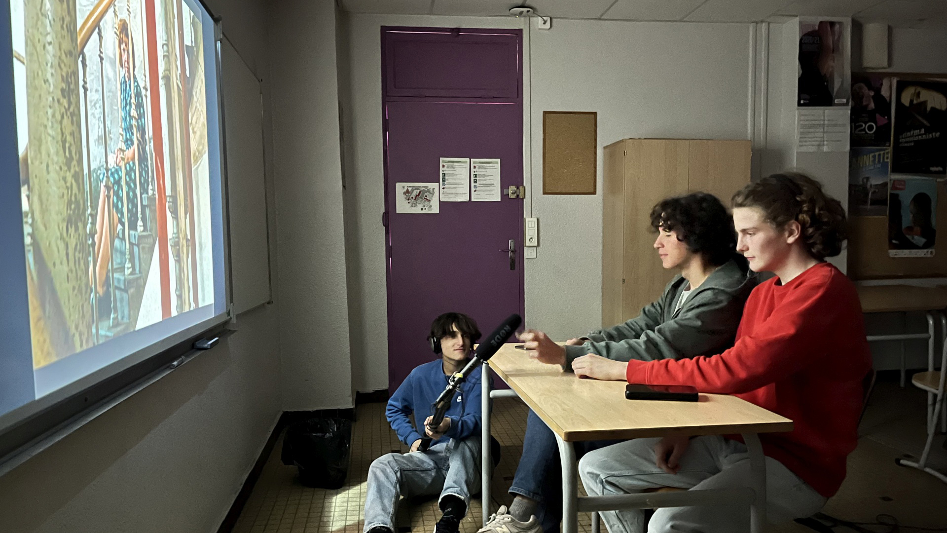 Zwei Jugendliche betrachten einen Film auf einer Leinwand, ein weiterer Jugendlicher hält ihnen ein Mikro entgegen
