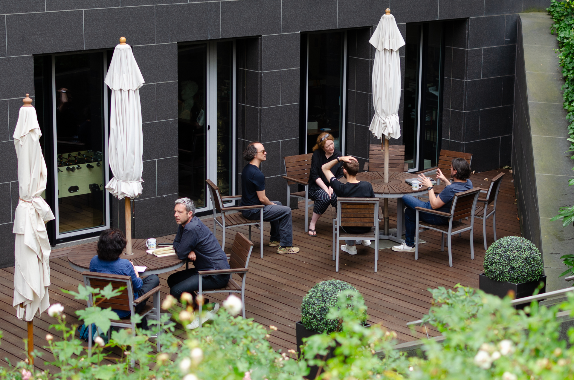 Menschen sitzen in Gruppen an Tischen auf einer Terrasse.