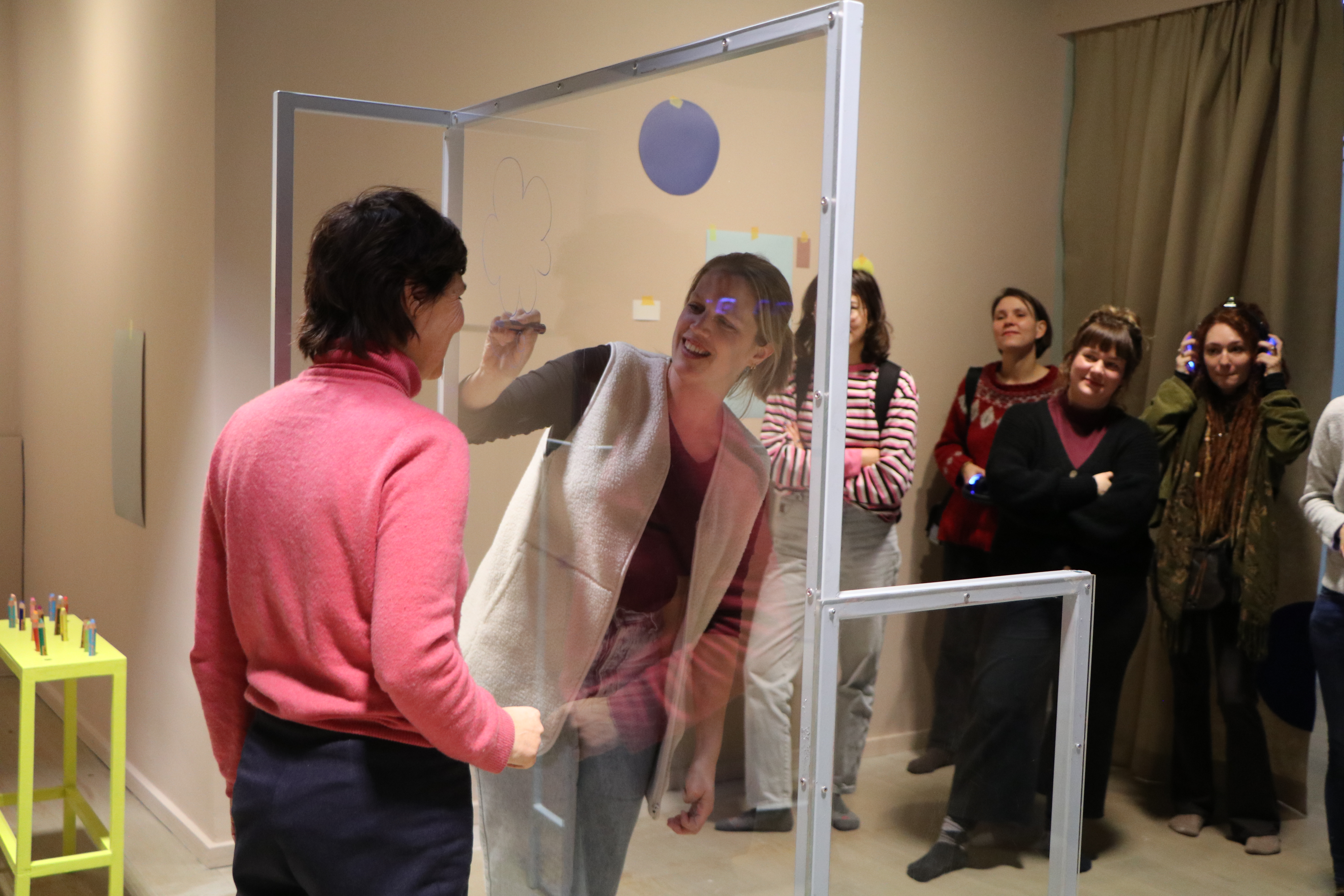 Zwei Personen stehen sich durch eine Plexiglasscheibe getrennt gegenüber, eine der beiden zeichnet etwas mit einem Stift auf die Scheibe. Im Hintergrund stehen Zuschauer:innen
