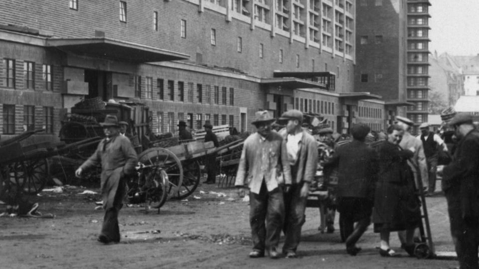 Eine Schwarzweißaufnahme einer Gruppe von Menschen, hauptsächlich Männer, die in verschiedenen Richtungen vor einem großen Gebäude entlanglaufen. Im Hintergrund stehen große Transportkarren.