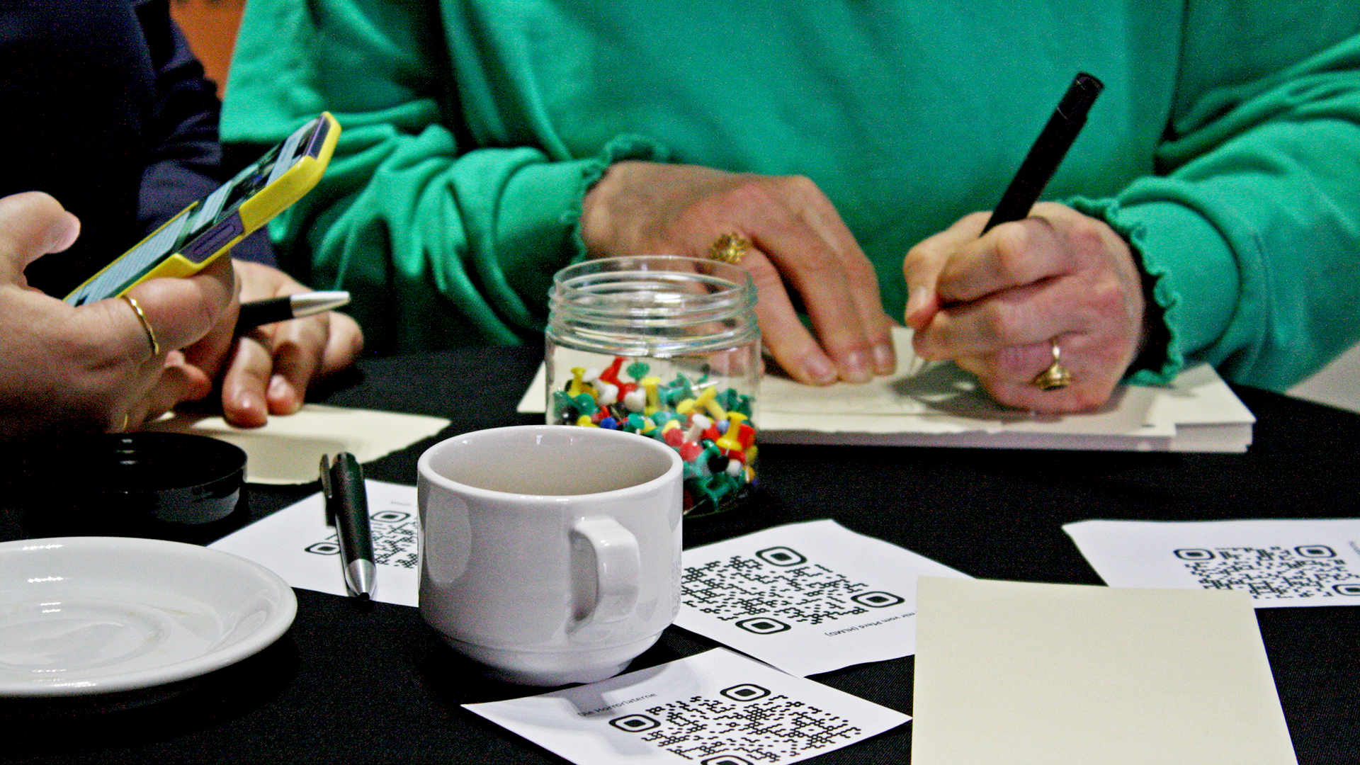 Zwei Handpaare, die Stifte und ein Smartphone halten. Eine Person schriebt etwas. Auf dem Tisch vor ihnen steht eine Tasse und liegen Pinnnadeln, Notizzettel und Zettel mit QR-Codes